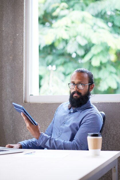 Indian man sitting at desk in office with tablet and looking towards camera