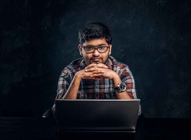 Indian programmer sits at a desk with a laptop and look at the camera with a thoughtful look. Studio photo against a dark textured wall.