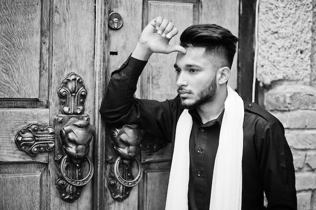 Indian stylish man in black traditional clothes with white scarf posed outdoor against old gates