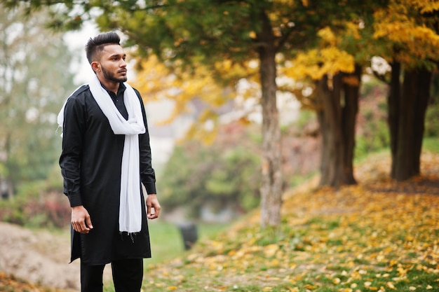 Indian stylish man in black traditional clothes with white scarf posed outdoor against yellow autumn leaves tree