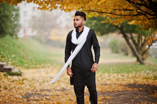 흰색 스카프를 두른 검은색 전통 옷을 입은 인도의 세련된 남자는 노란 단풍 나무에 야외 포즈를 취했습니다.