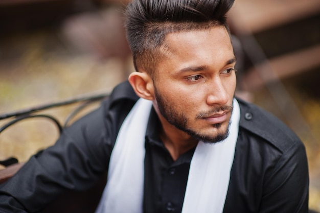 Indian stylish man in black traditional clothes with white scarf posed outdoor sitting on bench