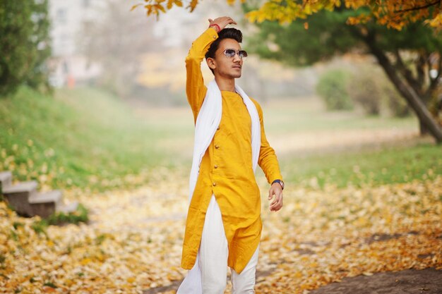 Индийский стильный мужчина в желтой традиционной одежде с белыми солнцезащитными очками-шарфами позирует на открытом воздухе на фоне осенних листьев дерева
