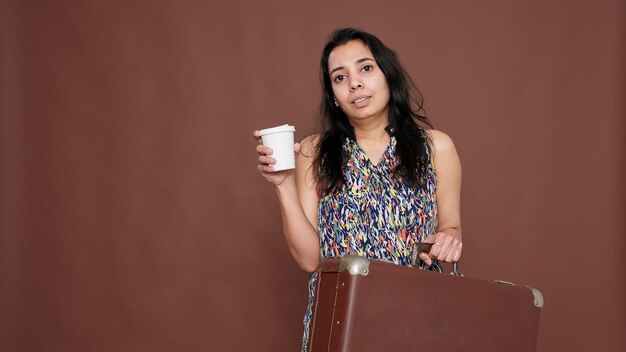 Indian woman getting ready to travel on vacation with baggage case and cup of coffee in hand. Feeling happy about leaving on holiday vacation trip with suitcase luggage and drink.