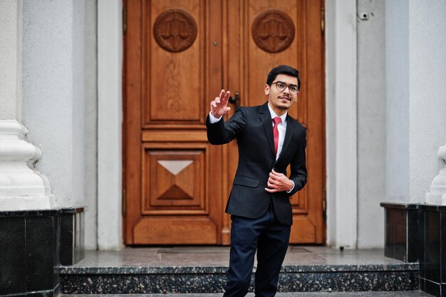 眼鏡をかけているインドの若い男は、建物のドアに対して屋外でポーズをとった赤いネクタイと黒のスーツを着て2本の指を示しています