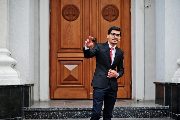 Индийский молодой человек в очках в черном костюме с красным галстуком стоит на улице у двери здания, показывает два пальца