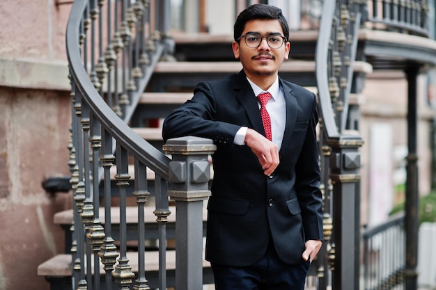 Indian young man at glasses wear on black suit with red tie posed outdoor against iron stairs