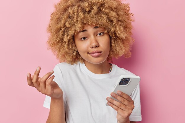 Indifferent careless woman shrugs shoulders feels hesitant has bored uninterested expression uses mobile phone for chatting online dressed in casual basic t shirt isolated over pink background