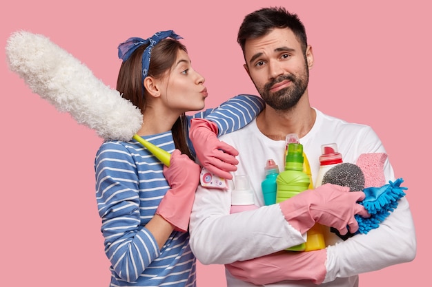 Free photo indifferent fatigue unshaven man holds many bottles of detergent, his wife leans at shoulder, wants to kiss in cheek, holds pp duster, ready to bring house in order and clean room carefully.