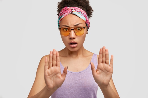 Бесплатное фото Возмущенная афроамериканка с недовольным выражением лица делает стоп-жест, что-то отвергает, носит модные оттенки и повседневный пурпурный жилет, изолированный на белой стене. прекрати это, пожалуйста