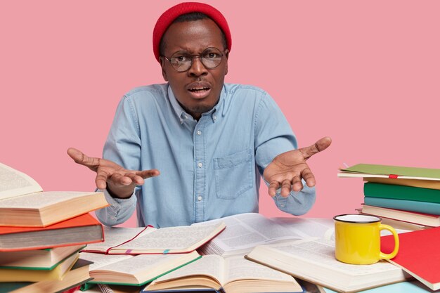 Indignant black teenage schoolboy gestures with bewilderment, wears red hat and formal shirt, asks what exactly he should do, reads textbooks