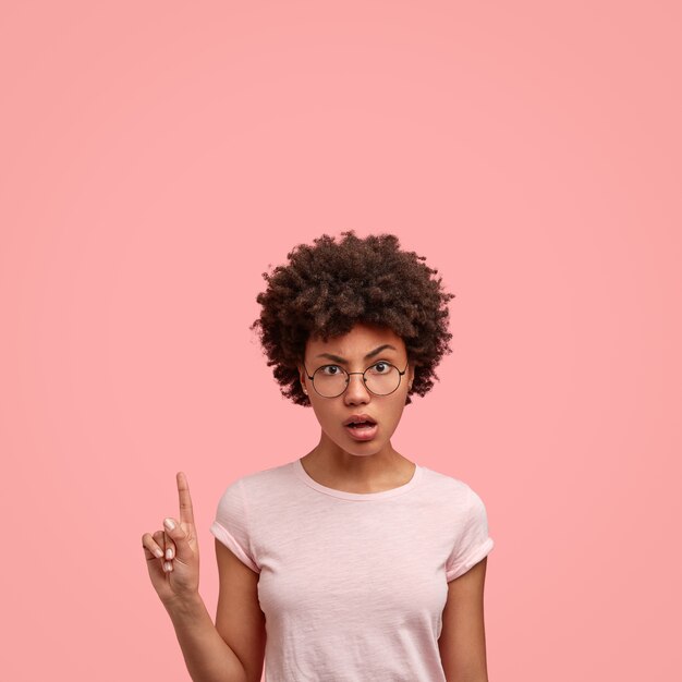 Indignant dark skinned curly lady points with index finger upwards