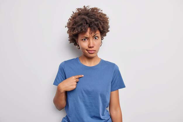 Indignant displeased curly haired young woman points at herself with angry expression asks who me wears casual blue t shirt puzzled being selected isolated over white background. Do you mean me