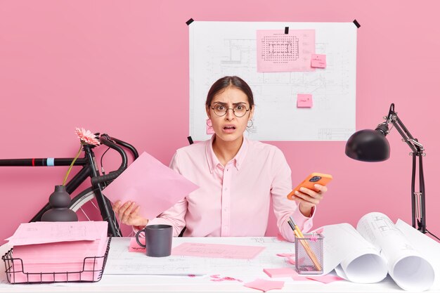 indignant displeased female office worker has much paper work feels confused holds mobile phone tries to generate ideas for engineering project poses in coworking space makes sketches blueprints