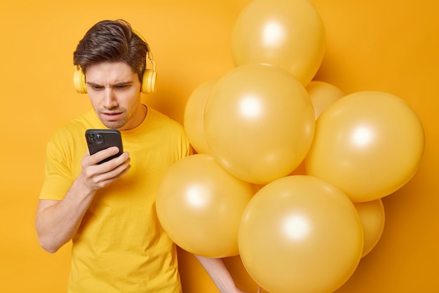 L'uomo dispiaciuto indignato concentrato sullo schermo dello smartphone legge notizie scioccanti vestito con una maglietta casual tiene un mucchio di palloncini gonfiati isolati su sfondo giallo tempo di festa e tecnologia