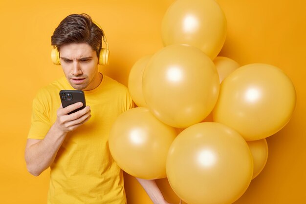 Indignant displeased man focused at smartphone screen reads shocking news dressed in casual t shirt holds bunch of inflated balloons isolated over yellow background Party time and technology