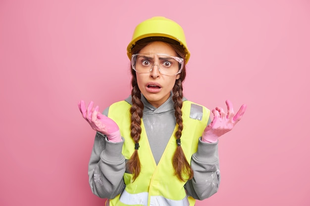 憤慨した不機嫌な女性ビルダー 産業労働者は肩をすくめ、困惑を感じ、手を横に広げ、保護用ヘルメット メガネを着用し、工場や建設現場で反射ベストをする