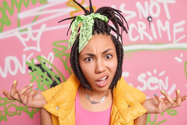 Indignant hipster girl has dreadlocks spreads palms says so what dressed in stylish clothes poses against colorful graffiti wall painted with aerosol spray