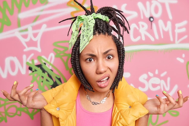 Indignant hipster girl has dreadlocks spreads palms says so what dressed in stylish clothes poses against colorful graffiti wall painted with aerosol spray