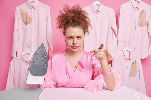 Indignant housewife with combed curly hair points  and reproaches you wears dressing gown holds steam electric iron busy ironing clothes for family. Laundry worker has angry expression