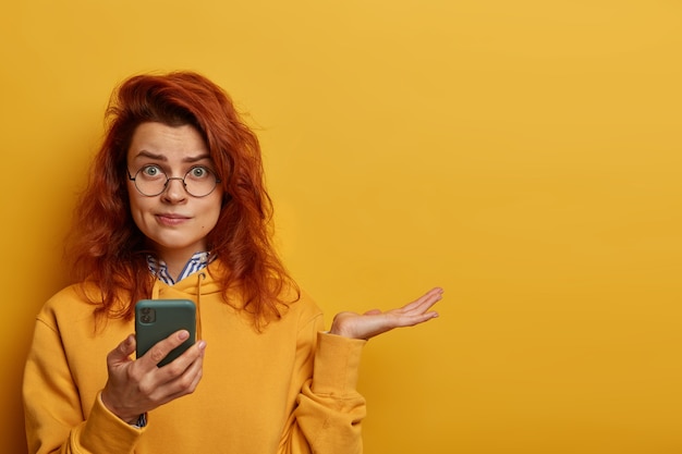 Indignant puzzled redhead woman raises palm, thinks what to answer on received message, holds mobile phone, wears round spectacles and hoodie, models over yellow wall with blank space right.