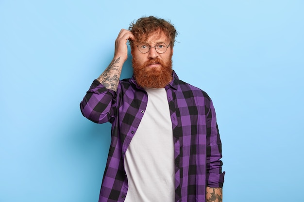 Indignant puzzled stylish ginger guy posing against the blue wall