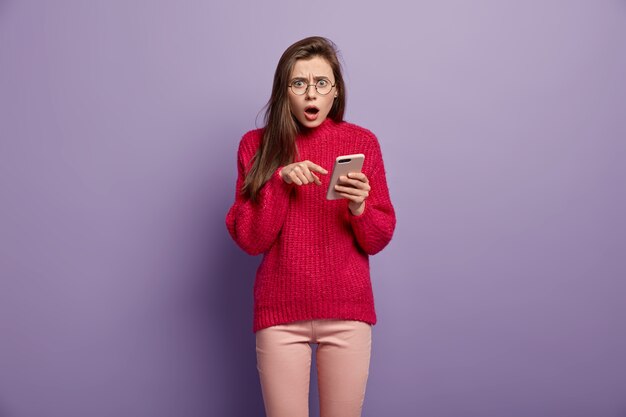 Возмущенная потрясенная молодая женщина показывает на мобильный телефон, демонстрирует что-то удивительное, удивлена полученным сообщением, носит красный свитер с длинными рукавами, у нее удивленное лицо, она подключена к Wi-Fi