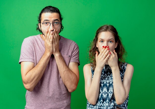 緑の壁の上に立っている手で口を覆うことにショックを受けて不快になっている憤慨している若いカップルの男性と女性