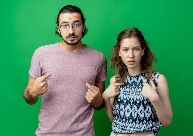 indignant young couple man and woman pointing at themselves standing next to each other over green wall
