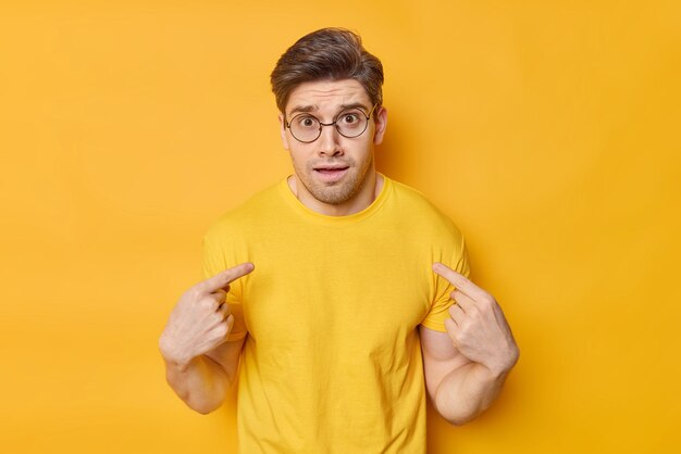 분개한 청년은 자신을 어리둥절해하며 투명한 둥근 안경을 쓰고 캐주얼한 티셔츠를 입고 선명한 노란색 배경에 고립된 나를 비난합니다.