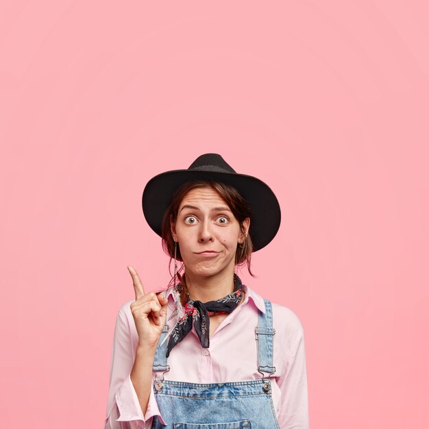 Indignant young woman florist in casual wear, poinst upwards with fore finger, has puzzled facial expression