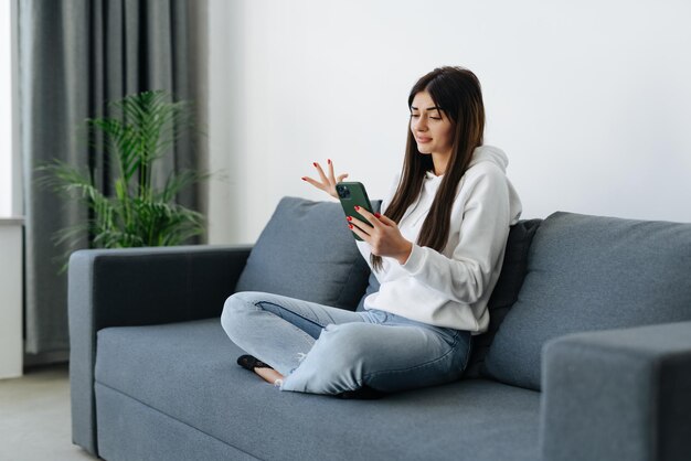 Indignant young woman sitting on couch in living room holding mobile phone having problems with digital device feels angry and annoyed