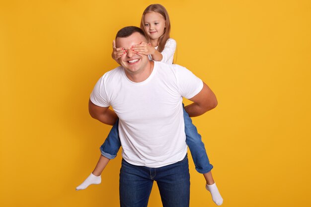 Indooor shot of happy father piggybacking daughter while charming kid closing his eyes, happy man with adorable girl