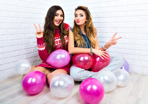 Indoor bright portrait of two funny bet friends sister hipster womans, going crazy, party time, pink balloon, showing v science, hugs and kisses, make up, sweaters, amazing smile.