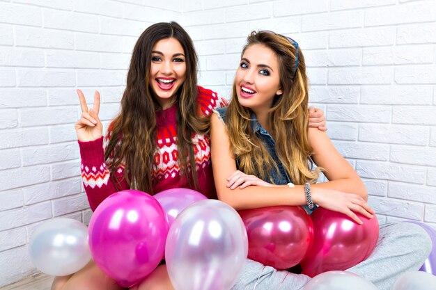 Indoor bright portrait of two funny bet friends sister hipster womans, going crazy, party time, pink balloon, showing v science, hugs and kisses, make up, sweaters, amazing smile.