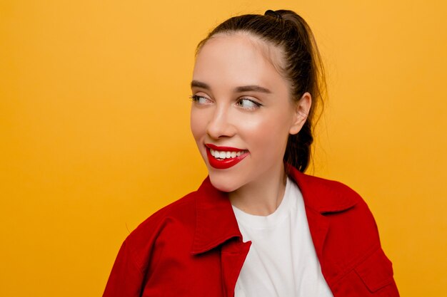 Indoor close-up portrait of beautiful female model with red lips, collected hair smiles and looking aside on isolated yellow wall, place for text