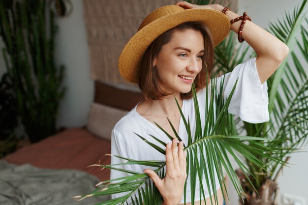 Indoor close up portrait of elegant pretty woman in straw hat and white blouse posing at home