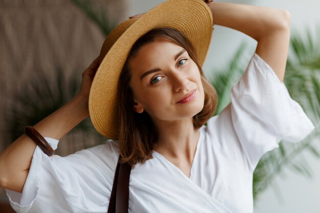 Indoor close up portrait of elegant pretty woman in straw hat and white dress posing at home