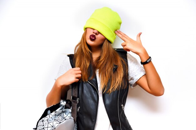 Indoor fashion grunge portrait of cheeky hipster woman ,leather jacket, rock style, dark lips, flash, crazy emotions. Put hat on her eyes, imitating gun by her hand, angry, evil.