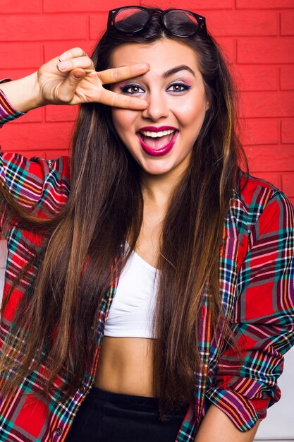 Indoor fashion lifestyle portrait of young pretty hipster woman with bright make up and amazing smile, have long brunette hairs, wearing plaid shirt. Red urban wall.