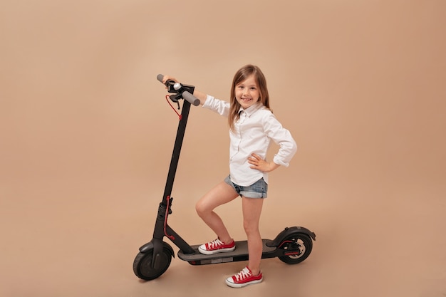 Indoor frame of little cute girl posing at camera over isolated beige background with electric scooter