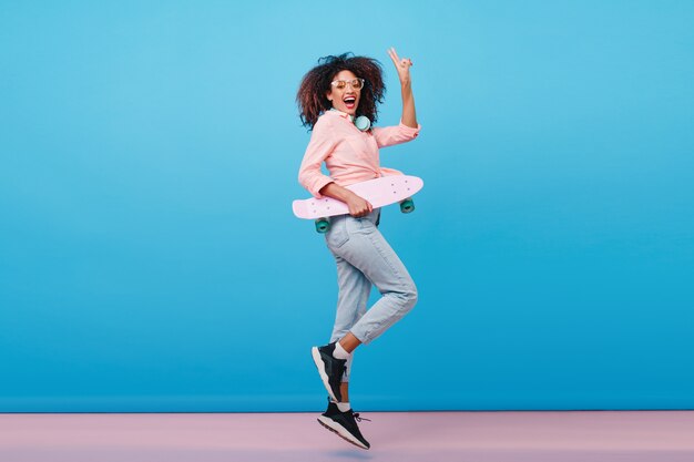 스케이트 보드를 들고 분홍색 셔츠에 자신감이 아프리카 여자의 실내 전신 초상화. 블루 인테리어와 함께 포즈 곱슬 헤어 스타일으로 열정적 인 흑인 여성.