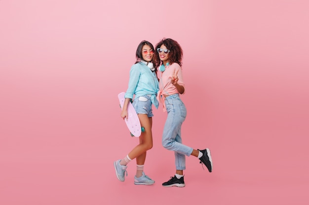 Indoor full-length portrait of sporty african girl in black shoes spending time with friend. Ecstatic asian lady with longboard standing near mulatto woman.