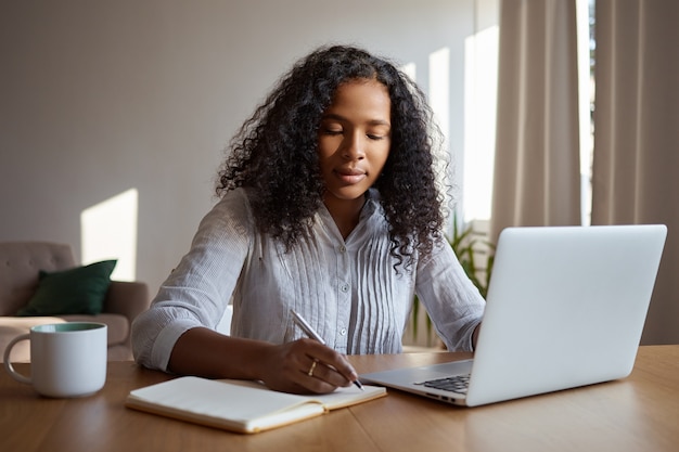 Indoor image of beautiful young dark skinned female with curly hair writing down in copybook making plans for day while sitting at desk with cup of coffee in front of open portable computer