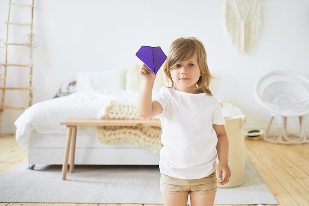 Indoor image of charming little European girl in casual clothes is playing indoors, holding with violet paper plane. Children, fun, games, activity and leisure time