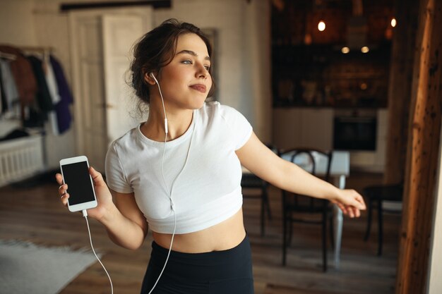 Indoor image of fashionable pretty girl wearing tight fitting top, moving body to rhythm of music, using mobile phone and earphones.