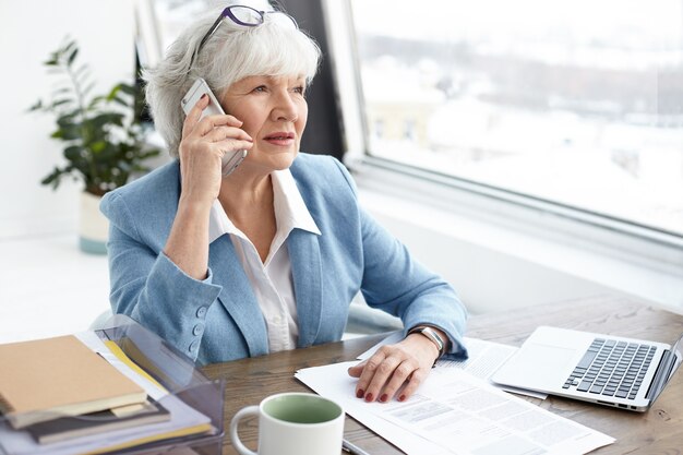 세련된 사무실에서 일하는 회색 머리 60 세 성숙한 여성 은행원의 실내 이미지, 휴대 전화에서 그녀의 고객과 민사 사건의 세부 사항을 논의하고, 노트북을 사용하여 창으로 책상에 앉아