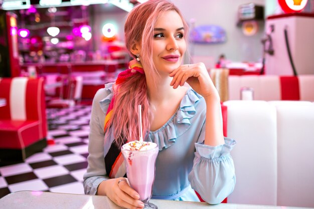 Indoor image of pretty young elegant woman enjoying her tasty sweet strawberry milk shake at retro vintage American restaurant, neon design, cute pastel dress, pink hairs and accessories