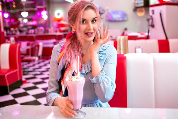 Indoor image of pretty young elegant woman enjoying her tasty sweet strawberry milk shake at retro vintage American restaurant, neon design, cute pastel dress, pink hairs and accessories