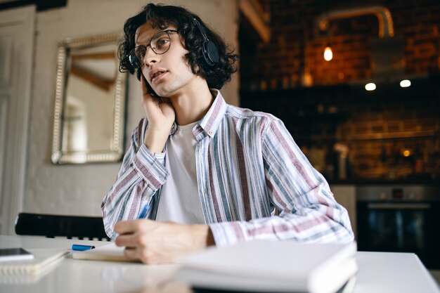 Indoor image of serious young male with curly hair sitting at his workplace with textbooks, writing down while listening to lecture via wireless headphones, learning from home. Social distancing